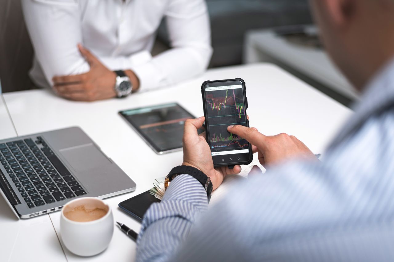 about-01 Two businessmen analyze stock market data using a smartphone and laptop in a modern office setting.